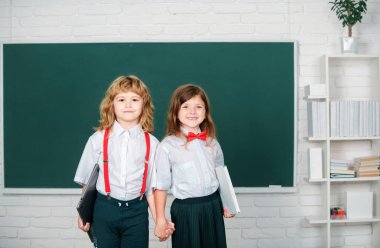 Portrait of two happy school kids studying in classroom at elementary school. Concept of friendship and classmate. Best school friends holding hand near blackboard