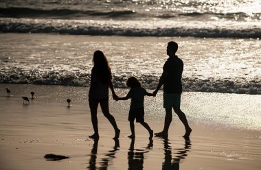 Sahilde yürüyen aile silueti. Anne, baba ve çocuk altın kumda yürüyor, anne ve baba kızların elini tutuyor. Tam boy. Aile açık hava aktiviteleri.