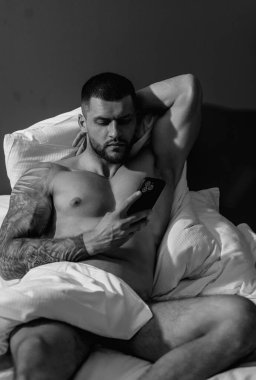 Akıllı telefon kullanarak yatağa uzanan seksi adam. Seksi adamın kaslı vücudu yatakta. Güçlü, vahşi adam. Seksi çıplak erkek gövdesi. Yatakta dövmeli çıplak kaslı bir adam yatıyordu. Yatakta yatan kaslı adam.