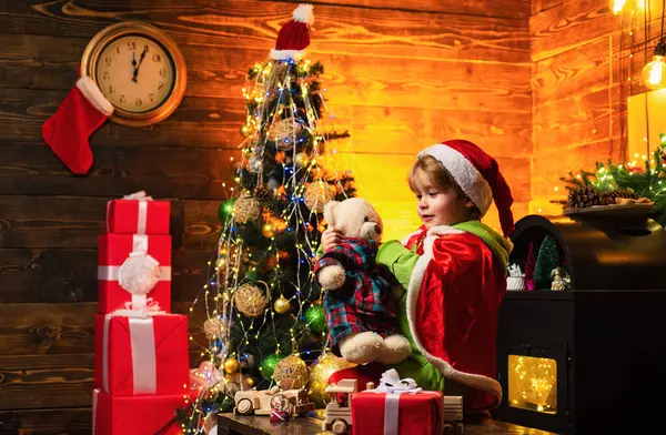 Mutlu çocuk iki eliyle kırmızı bir hediye kutusu tutuyor. Noel kutlaması tatili. Kışın giyinmiş küçük mutlu çocuk Noel ağacının yanında Noel Baba 'yı düşün.