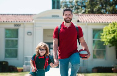 İlkokulun ebeveynleri ve öğrencileri el ele gider. Tişörtlü öğretmen ve okul parkının yanında sırt çantalı sevimli okul çocuğu.