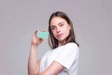 Yüzünün yanında kredi kartı tutan beyaz tişörtlü genç bir kadın. Gri arka planda izole edilmiş. Zeka ya da banka kartını öde. Banka kartı. Plastik kredi kartı