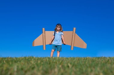Child boy plays in an astronaut dreams of space. Happy child play with toy plane cardboard wings against blue sky. Kid having fun in summer field outdoor. Portrait of boy with paper wings