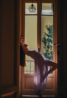 Uzun siyah saçlı güzel bir genç kadın. Rahatlayan güzel, gülümseyen bir kadın. - Günaydın. Şehvetli model kız. Şehvetli kadın. Kadın sabah gündoğumuyla uyandıktan sonra pencerenin yanında duruyor.