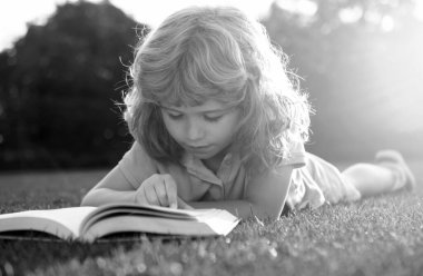 Kid boy reading a book lying on grass. Cute little child in casual clothes reading a book and smiling while lying on grass in park. Smart clever Kids