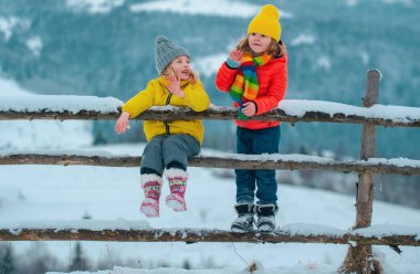 İki küçük çocuk, karla kaplı kırsal dağlarla güzel kış doğasında eğleniyor. Çocuklar için kışın örülmüş kıyafetler.