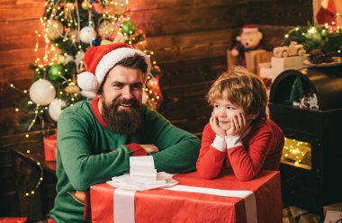 . Kış kıyafetleri içinde babası olan mutlu bir çocuk Noel ağacının yanında Noel Baba 'yı düşün. Noel ailesi. Baba ve oğlu Noel hediyesi ile ahşap ev geçmişi hakkında