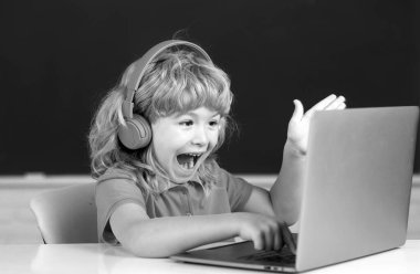 Heyecanlı kulaklıklı çocuk, masada oturur, okulda dizüstü bilgisayarda çalışır, klever şaşkın çocuk internet derslerini kullanarak not defterine kulaklık takar.