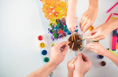 Anne, baba ve çocukların elleri renkli kalemlerle resim çiziyor. Resim malzemeleriyle beyaz masaya üst görünüm