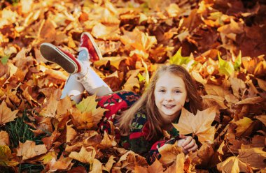 Yaprakların üzerinde yatan kız çocuğu. Güzellik parkında sonbahar yaprakları olan sevimli küçük bir kız. Sonbahar parkındaki komik çocuklar. Sonbahar portakal yapraklı küçük kız