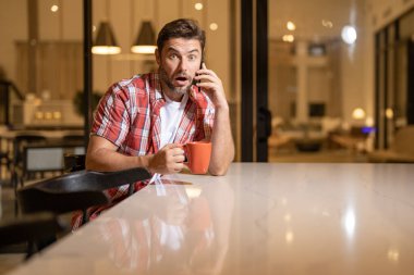 Oturma odasında akıllı telefona bakan adamın portresi. Adam elinde bir fincan kahveyle akıllı telefondan konuşuyor. Evde cep telefonu kullanan düşünceli bir adam. Telefon kullanan orta yaşlı bir adam.