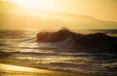 Dalgalar sıçrıyor. Altın gökyüzü ve deniz suyu. Günbatımında deniz manzarası ve tropikal plaj ya da tatil ve tatil için gün doğumu. Okyanus dalgaları arka planı. Suda güneş, kumsalda kum.