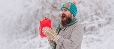 Örgü şapkalı ve kışlık süveterli heyecanlı bir adamın portresi. Noel hediyeli mutlu adam dışarıda. Karlı kış arkaplanı. Mutlu Noel Baba 'nın dışarıda kar yağarken çekilmiş fotoğrafı. Elinde hediyeler var.