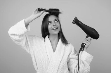 Bornozlu kadın saç tarıyor, saç kurutma makinesiyle saç kurutuyor. Saçlarını tarayan kadın modelin portresi. Saç fırçası ve saç kurutma makinesi olan kız. Saç bakımı ve güzellik. Sabah rutini