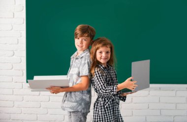 Okulda dizüstü bilgisayarı ve kitabı olan kız ve erkek çocukların portresi. Sınıftaki çocuklar tahtada