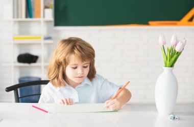 Öğrenci evde ders veriyor. Okullu çocuk bir şeyler yazıyor ve sınıftaki masada oturuyor. Birinci sınıf öğrencisi