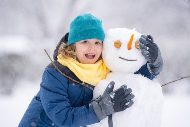 Soğuk bir kış gününde kış parkında bir çocuk. Kışın karla oynayan sevimli bir çocuk. Kardan adamla komik çocuk portresi. Noel 'de kış aktiviteleri. Karlı parkta kardan adam yapan çocuk