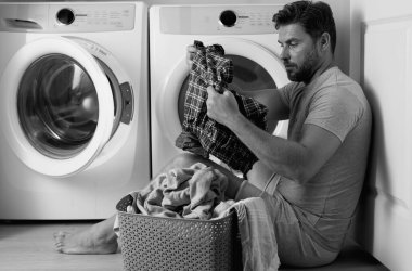 Çamaşır makinesinin yanında elbiseli bir adam. Yakışıklı adam çamaşır makinesinin önünde oturur. Çamaşır makinesini kirli çamaşırlarla dolduruyor. Adam elbiseleri temizliyor. Yalnız bir adam için ev işi. Ev çamaşırları