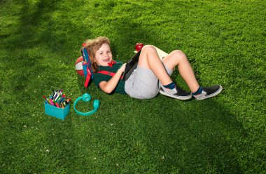 Parktaki çimde tabletle ve okul malzemeleriyle oturan bir çocuk. Açık hava okulu