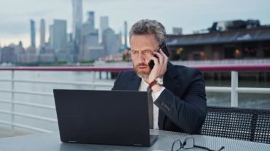 Bilgisayarda çalışan ve dışarıda telefonla konuşan bir iş adamı. İş konuşmaları. Meşgul iş adamı telefonda konuşuyor. Adam iş e-postası ve sörf yapmak için akıllı telefon kullanıyor.