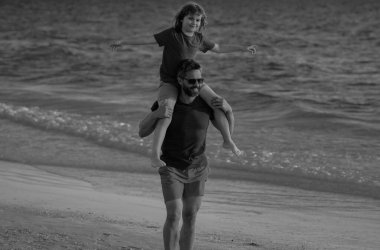 Oğlu babasının omuzlarına biniyor. Baba ve oğul sahilde yürüyor. Yaz tatili. Baba ve oğul birlikte yaz tatilinin tadını çıkarıyorlar. Baba oğul sahilde oynuyorlar.