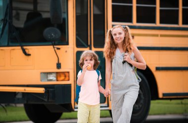 Okul otobüsündeki ilkokul öğrencileri. Çalışmaya biraz hazır. Mutlu küçük kız ve erkek kardeş okul otobüsünün önünde yan yana duruyorlar.