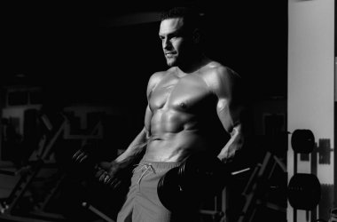 Spor salonunda pazı kaldıran bir sporcu. Dambılı bir sporcunun portresi. Gövdesi kaslı, tişörtsüz bir adam spor salonunda.