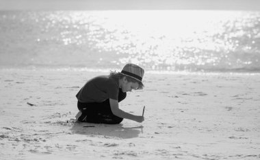 Şapkalı ve gömlekli sarışın çocuk deniz kenarındaki kuma resim çiziyor. Çocuklarla seyahat ve dinlenme. Çocukların mutluluğu. Kumda çizim yapan çocuk yaz kumsalında tek başına oynuyor.