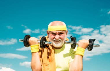 Dambıl kaldırarak egzersiz yapan mutlu bir son sınıf öğrencisi. Mavi gökyüzü arka planında büyük baba sportmen portresi. Spor merkezinde kıdemli sporcu.
