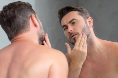 Bin yıllık İspanyol adam aynaya, yüz derisine ve sakalına bakıyor. Erkek güzellik ürünleri. Cilt bakımı, spa. Güzel bir adamın güzel portresi. Spa modeli, nemlendirici krem