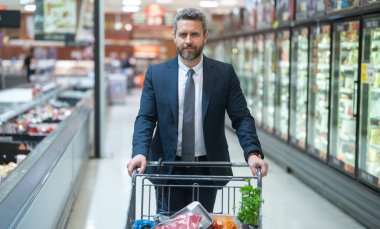 Markette takım elbiseli bir adam alışveriş yapıyor. Alışveriş arabalı bir işadamı market alışverişi yapıyor. Adam alışveriş arabasını marketin koridorlarında itiyor. Bakkalda alışveriş yapan bir adam.