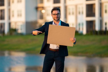 Açık havada poz veren boş afişi olan bir iş adamı. Takım elbiseli çekici bir adamın portresi boş boş bir poster tutuyor. Boş poster gösteren, işaret eden ve işaret eden bir adam.