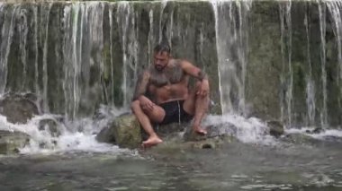 Şelaledeki seksi adam. Spor adamı doğada. İnsan özgürlüğü yaşam tarzı. Yaşam tarzı doğa, yazları doğa. Sakinlik ve doğada rahatlama. Güçlü gövdeli güçlü adam, dışarıda banyo yapmaktan hoşlanıyor.