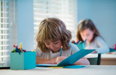 Sınıfta güzel çocuk çiziyor. Kitapla yazı yazan ve sınıftaki masada oturan okul çocuğu.