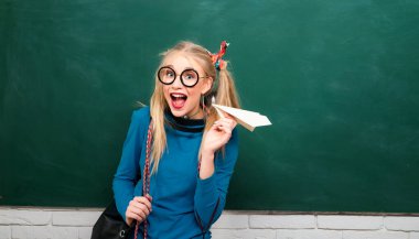 Young female high school student with paper plane in class. Funny girl in glasses. Charming girl in school is playing with paper plane and smiling. University student posing on blackboard clipart