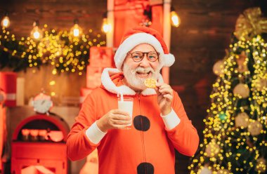 Şükran Günü ve Noel. Neşeli Noel Baba elinde sütle bardak, şömineli kurabiye ve arka planda Noel ağacı tutuyor.