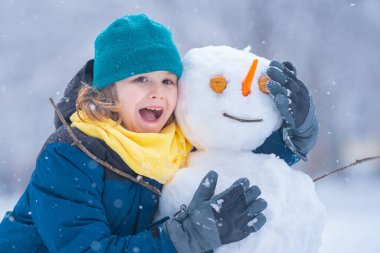 Soğuk bir kış gününde, kış parkında heyecanlı bir çocuk. Kışın karla oynayan sevimli bir çocuk. Kardan adamla komik çocuk portresi. Noel 'de kış aktiviteleri. Parkta kardan adam yapan çocuk