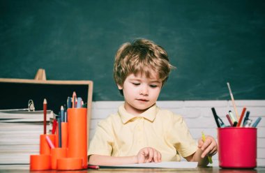 Kitap ve çanta ile İlköğretim Okulu çocuk. Çocuk arka planda bir tahta ile cevap vermeye hazırdır. Mutlu sevimli çalışkan çocuk kapalı bir masada oturuyor