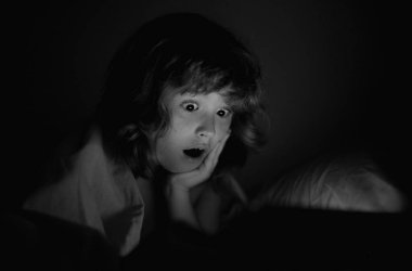 Dijital tablette oynayan bir çocuk. Çocuk oyun oynamak için tablet kullanıyor ve yatakta yatarken internetten öğreniyor. Yüzünde ekran ışığı refleksi var. Geceleri karanlık bir odada battaniyenin altında çocuk tableti oynuyor.