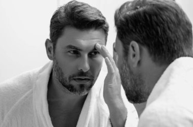 Bin yıllık İspanyol adam aynaya, yüz derisine ve sakalına bakıyor. Erkek güzellik ürünleri. Cilt bakımı, spa. Güzel bir adamın güzel portresi. Spa modeli, nemlendirici krem