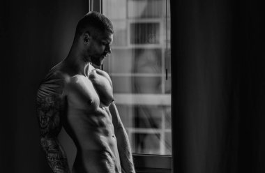 Man standing near window. Seductive gay. Muscular body of man in hotel room. Sexy guy with naked body. Nude muscular man body with tattoo. Sexy man with muscular chest in bedroom on window curtains