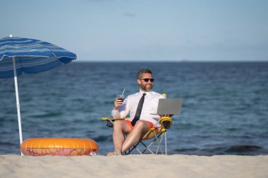Yaz tatillerinde başarılı bir iş adamı. Komik iş adamı deniz sahilinde yaz kokteyli içip dizüstü bilgisayar kullanıyor. Yaz işi konsepti. Sıcak yaz. Çılgınca. Uzak çevrimiçi çalışma