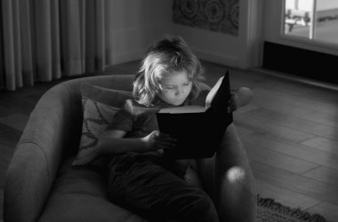 İlginç çocuk kitapları okuyan sevimli sarışın çocuk portresi. Odadaki kanepede uzanmış kitap okuyan sıradan giyinmiş tatlı bir çocuk.