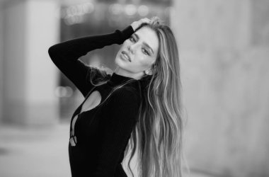 Uzun sarı saçlı, güzel, şehvetli bir kadın. Sokakta seksi bir manken. Açık havada siyah elbiseli bir manken. Güzel kadın açık hava portresi