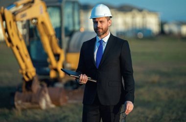İnşaat işçisi yatırımcı. İnşaat alanının önündeki yatırımcı. Modern ev inşaatında başarılı yakışıklı bir adam. Takım elbiseli ve kasklı orta yaşlı yatırımcının portresi
