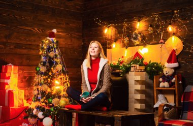 Elinde bir bardak sıcak içecekle yeni yılı bekleyen bir kadın. Mutlu genç bayan Noel ağacının yanındaki şöminenin yanında.