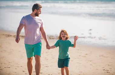 Baba ve oğul yaz plajında yürüyor. Baba ve çocuk el ele tutuşup birlikte yürüyorlar. Aile seyahati, tatil, babalar günü konsepti