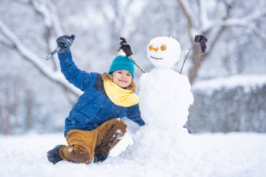 Soğuk bir kış gününde, kış parkında eğlenceli çocuk. Kışın karla oynayan sevimli bir çocuk. Kardan adamla komik çocuk portresi. Noel 'de kış aktiviteleri. Karlı parkta kardan adam yapan çocuk