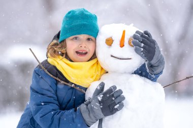 Soğuk bir kış gününde, kış parkında eğlenceli çocuk. Kışın karla oynayan sevimli bir çocuk. Kardan adamla komik çocuk portresi. Noel 'de kış aktiviteleri. Karlı parkta kardan adam yapan çocuk