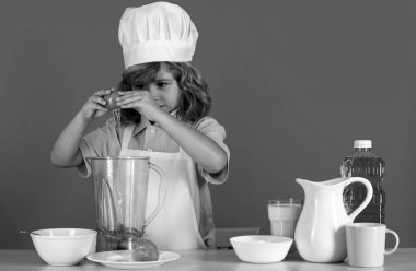 Çocuk aşçı, izole edilmiş mavi stüdyoda yemek hazırlıyor. Çocuklar yemek yapıyor. Önlüklü ve şapkalı genç çocuk mutfakta sağlıklı bir sebze yemeği hazırlıyor.